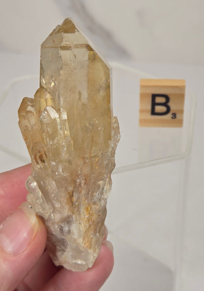 A hand holding a natural quartz crystal cluster with a small wooden letter tile labeled 'B3' in the background.