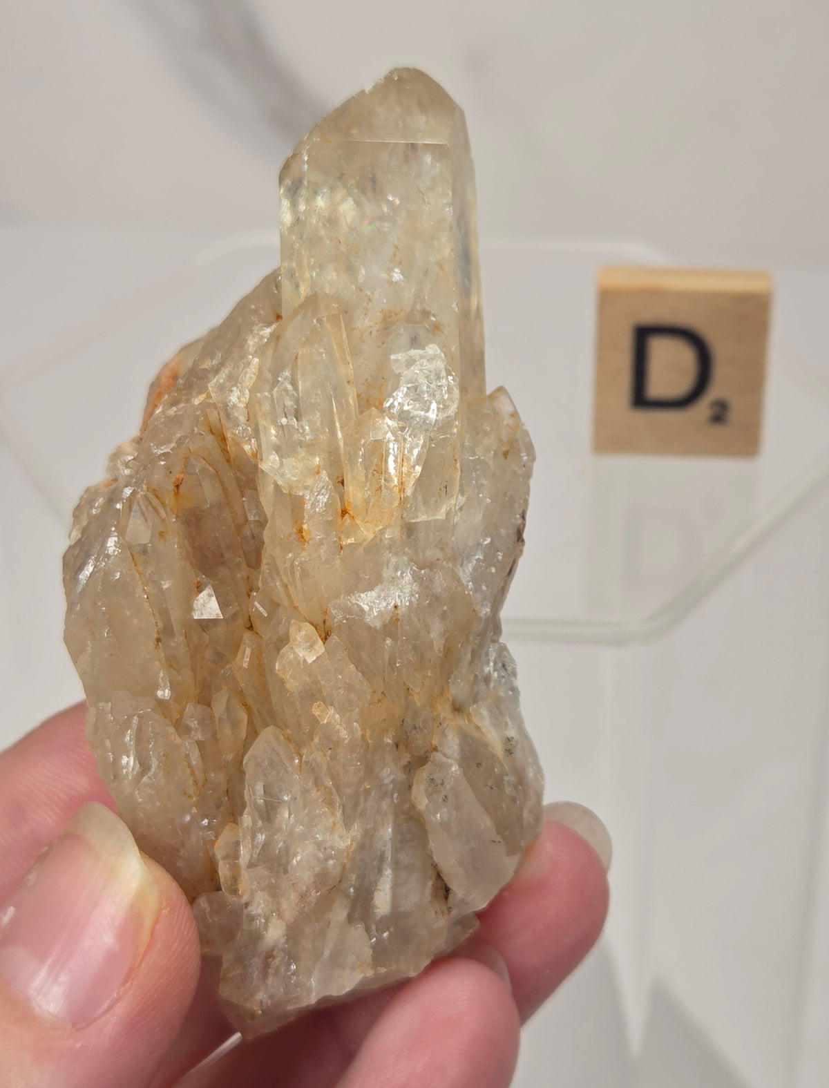 A hand holding a raw, natural clear quartz crystal cluster with orange-brown inclusions, with a small Scrabble tile showing the letter 'D' in the background.