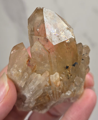 A hand holding a natural quartz crystal cluster with a prominent central clear and slightly smoky crystal point, set against a light gray marble background.