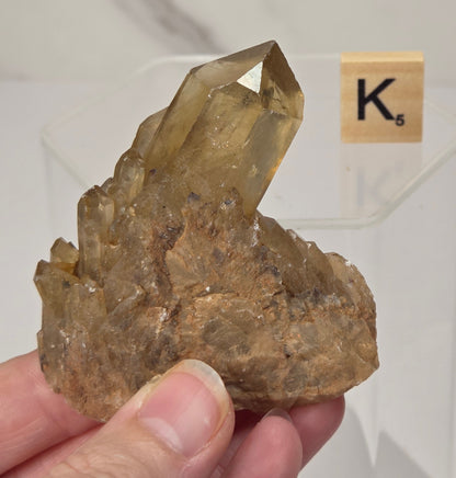 A hand holds a natural smoky quartz crystal cluster with a prominent pointed crystal tip. A small Scrabble tile spelling 'K' is visible in the background on a clear acrylic stand.