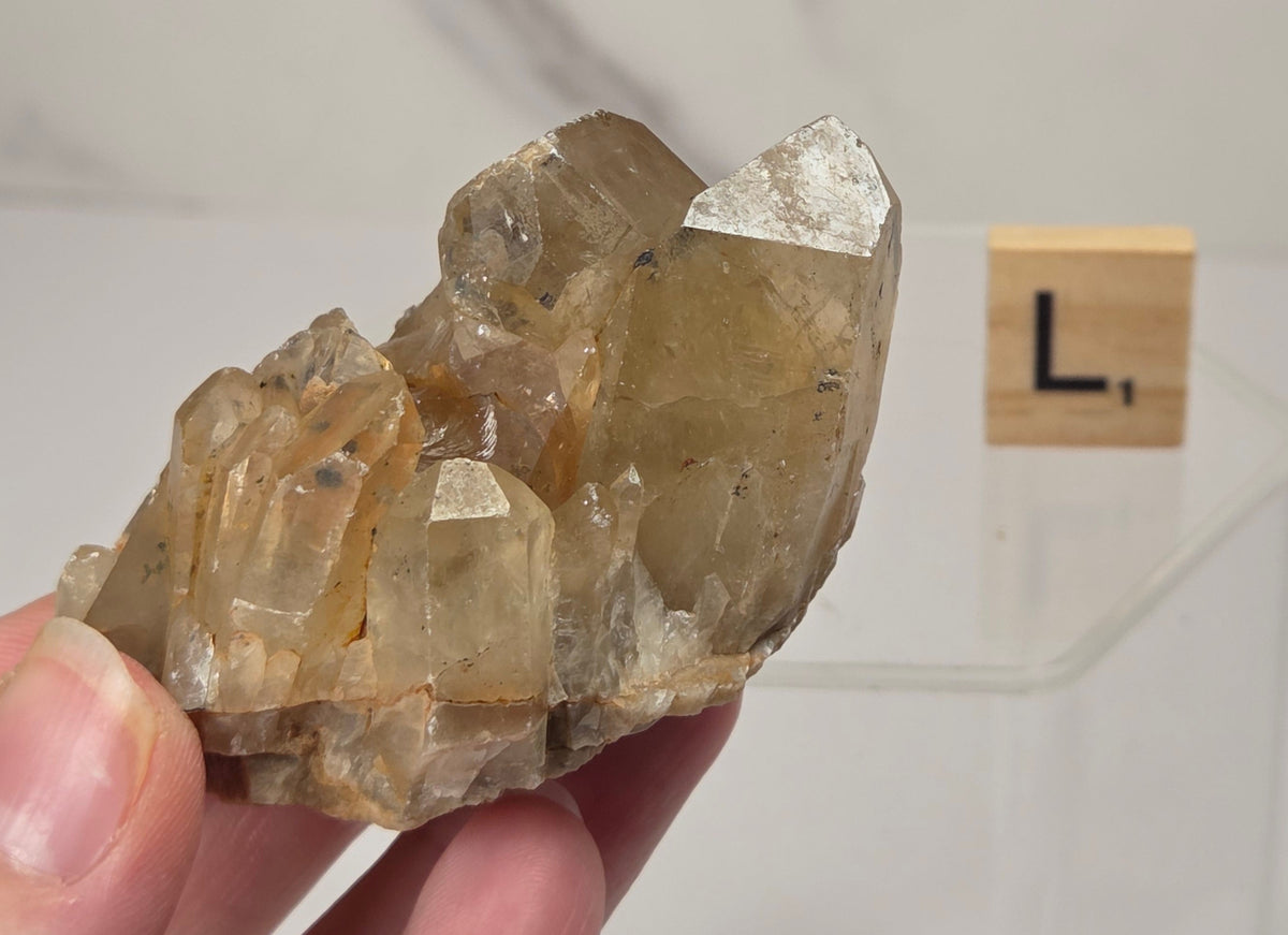 A hand holding a natural quartz crystal cluster with a metallic silver point, set against a clean white marble background with a small wooden letter block displaying 'L' in the distance.