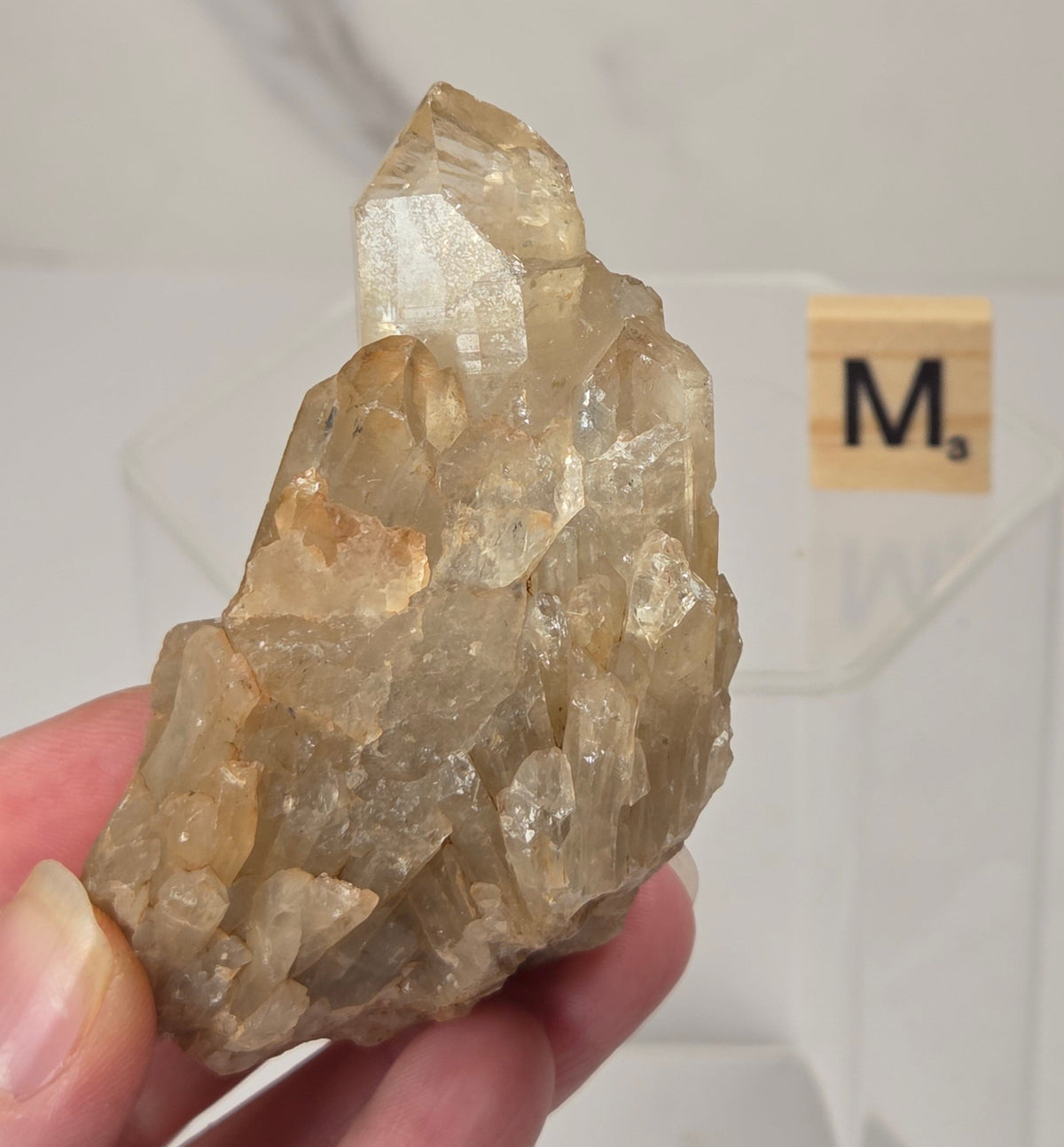 A hand holding a raw, rough quartz crystal specimen with natural facets and a warm tan-brown color, set against a light gray marble background with a clear acrylic display block.