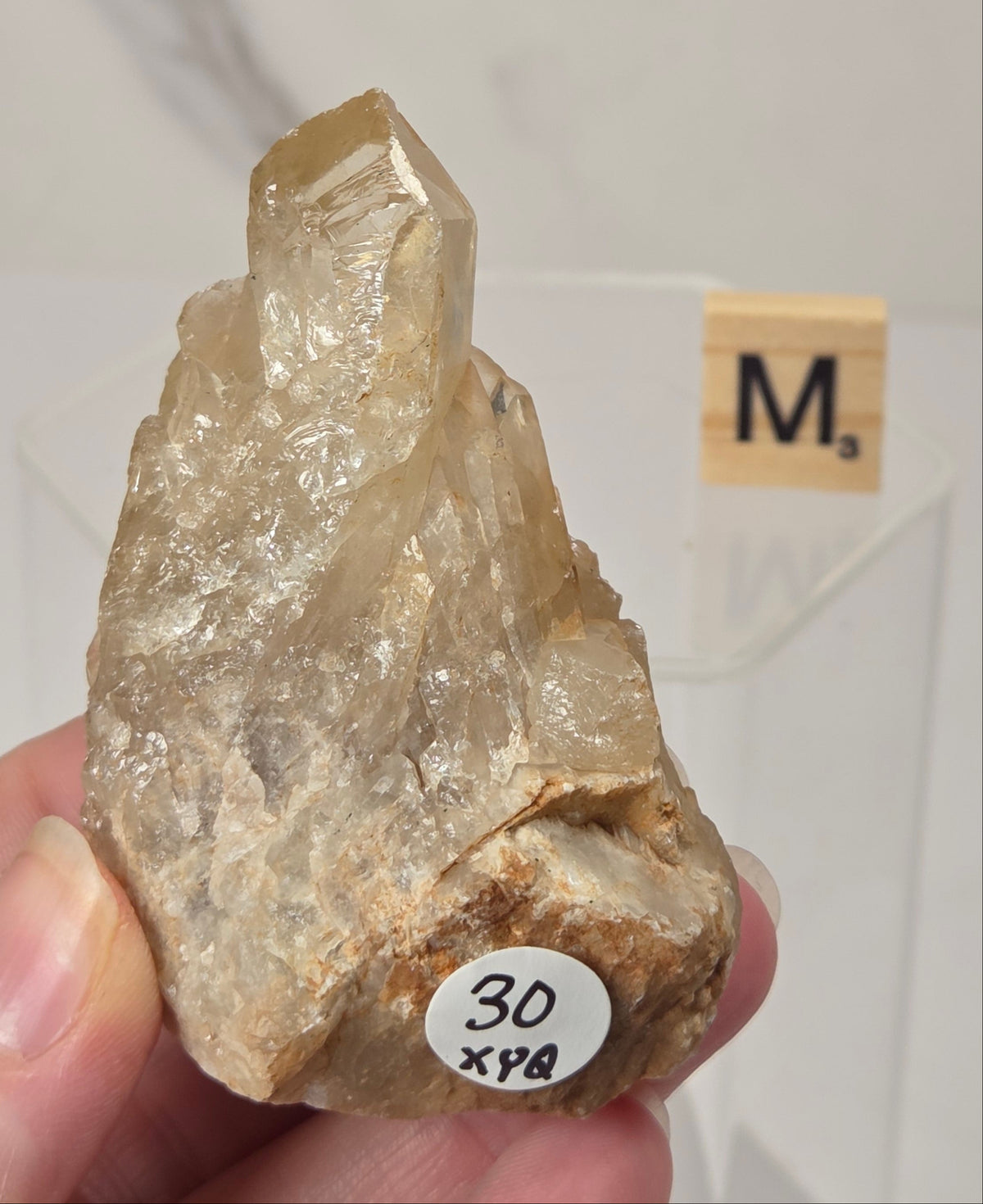 A hand holding a raw quartz crystal specimen with a rough matrix, featuring a small white sticker with '30 x98' and a small wooden tile with 'M' in the background.