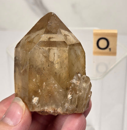 A hand holding a natural translucent quartz crystal cluster with a pointed top, shown against a light gray marble background with a small letter tile in the distance.
