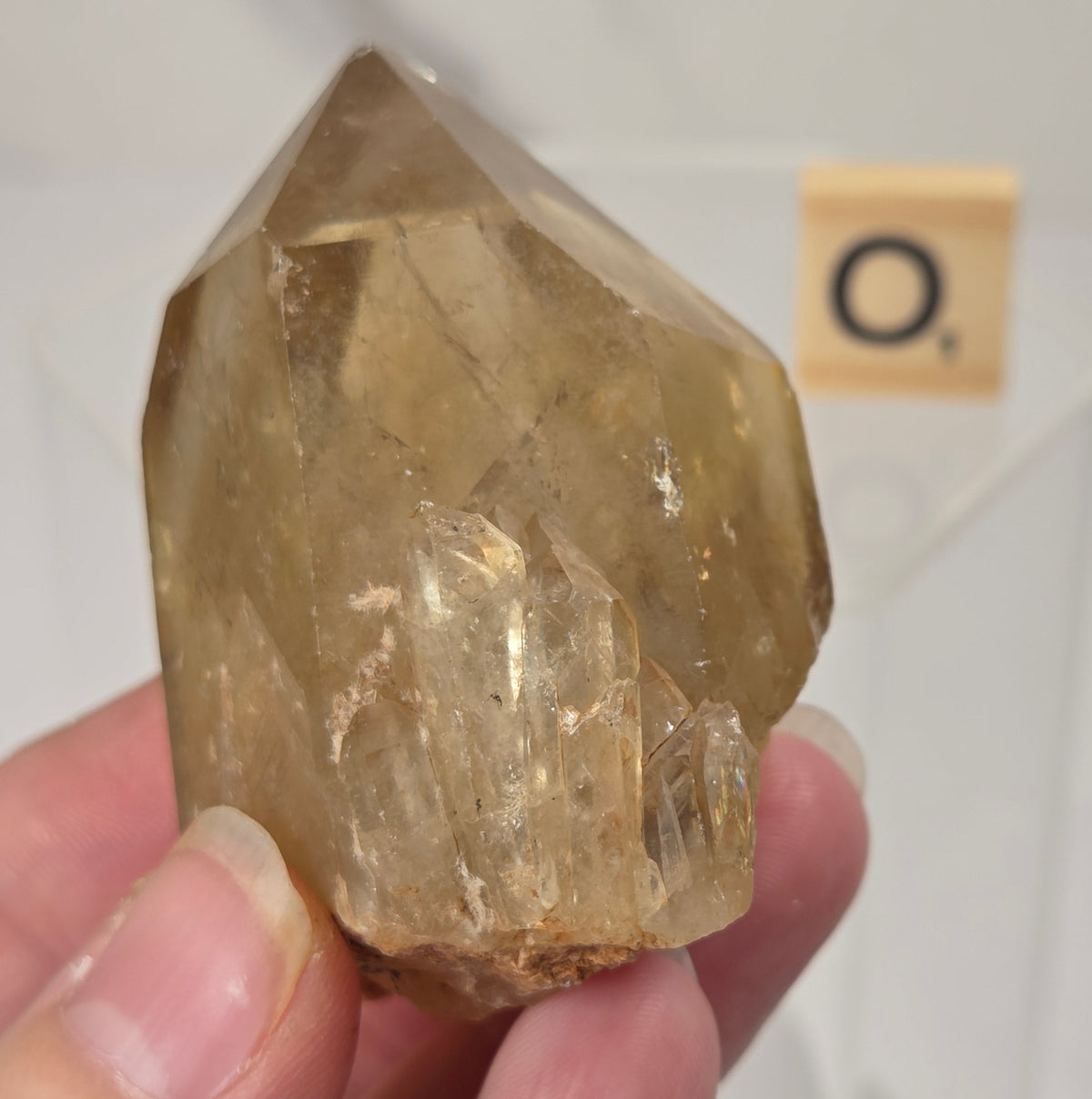 A hand holding a natural clear quartz crystal point with yellowish-brown inclusions, photographed against a white background with a small Scrabble tile in the distance.
