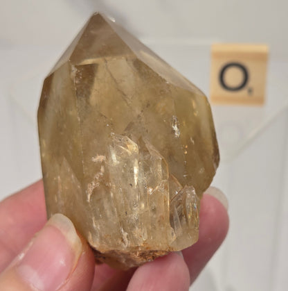 A hand holding a natural clear quartz crystal point with yellowish-brown inclusions, photographed against a white background with a small Scrabble tile in the distance.