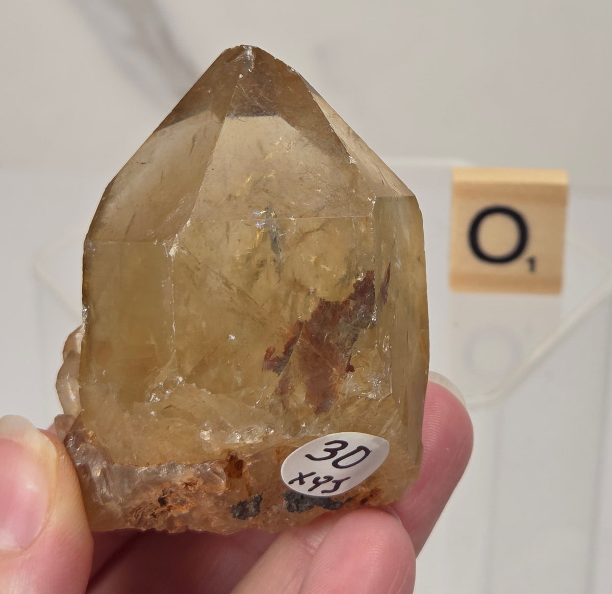 A hand holding a natural quartz crystal point specimen with a rough matrix base. The crystal is translucent golden-brown with some darker inclusions.