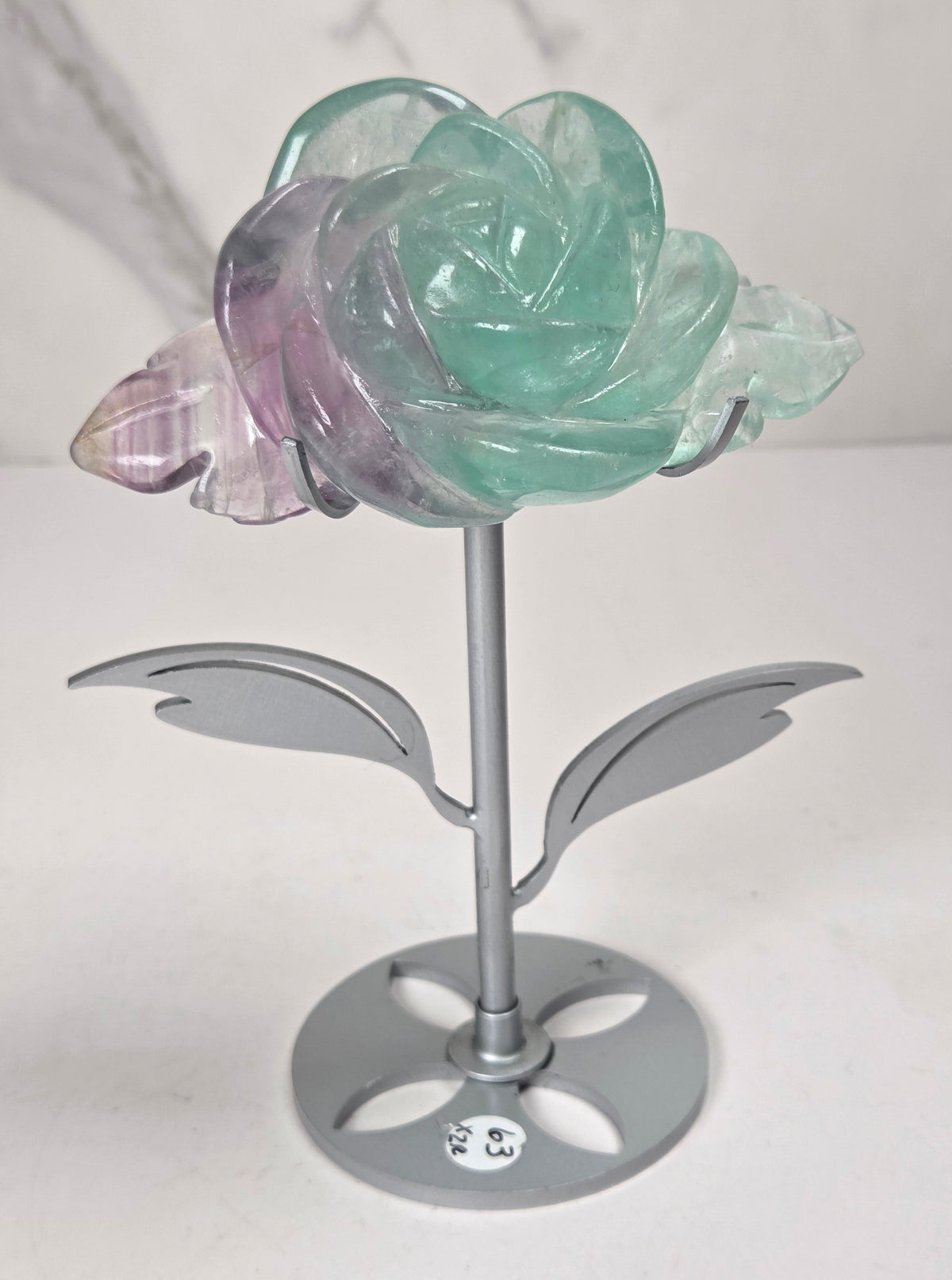 A decorative flower-shaped metal stand with a translucent rose head in mint green and lavender-purple tones, set against a light gray marble-patterned background.