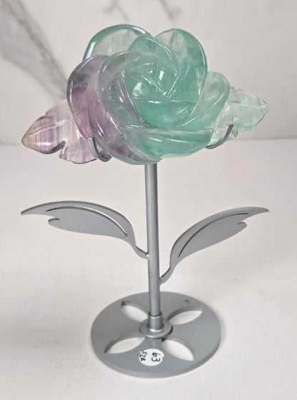 A decorative flower-shaped metal stand with a translucent rose head in mint green and lavender-purple tones, set against a light gray marble-patterned background.