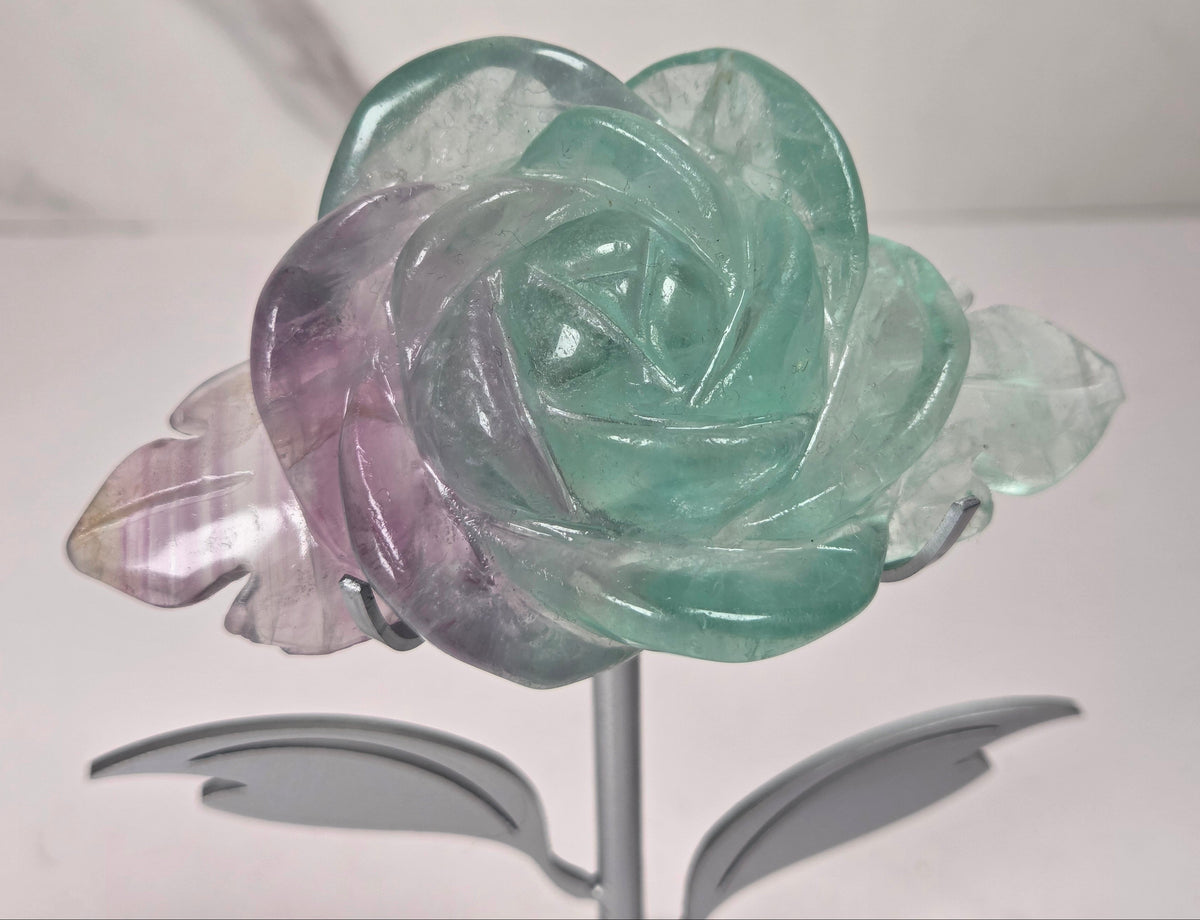 A carved rose-shaped decorative object made of translucent stone with a color gradient from light aqua blue to soft lavender-pink, mounted on a silver metal stand against a plain light background.