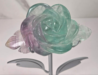 A carved rose-shaped decorative object made of translucent stone with a color gradient from light aqua blue to soft lavender-pink, mounted on a silver metal stand against a plain light background.
