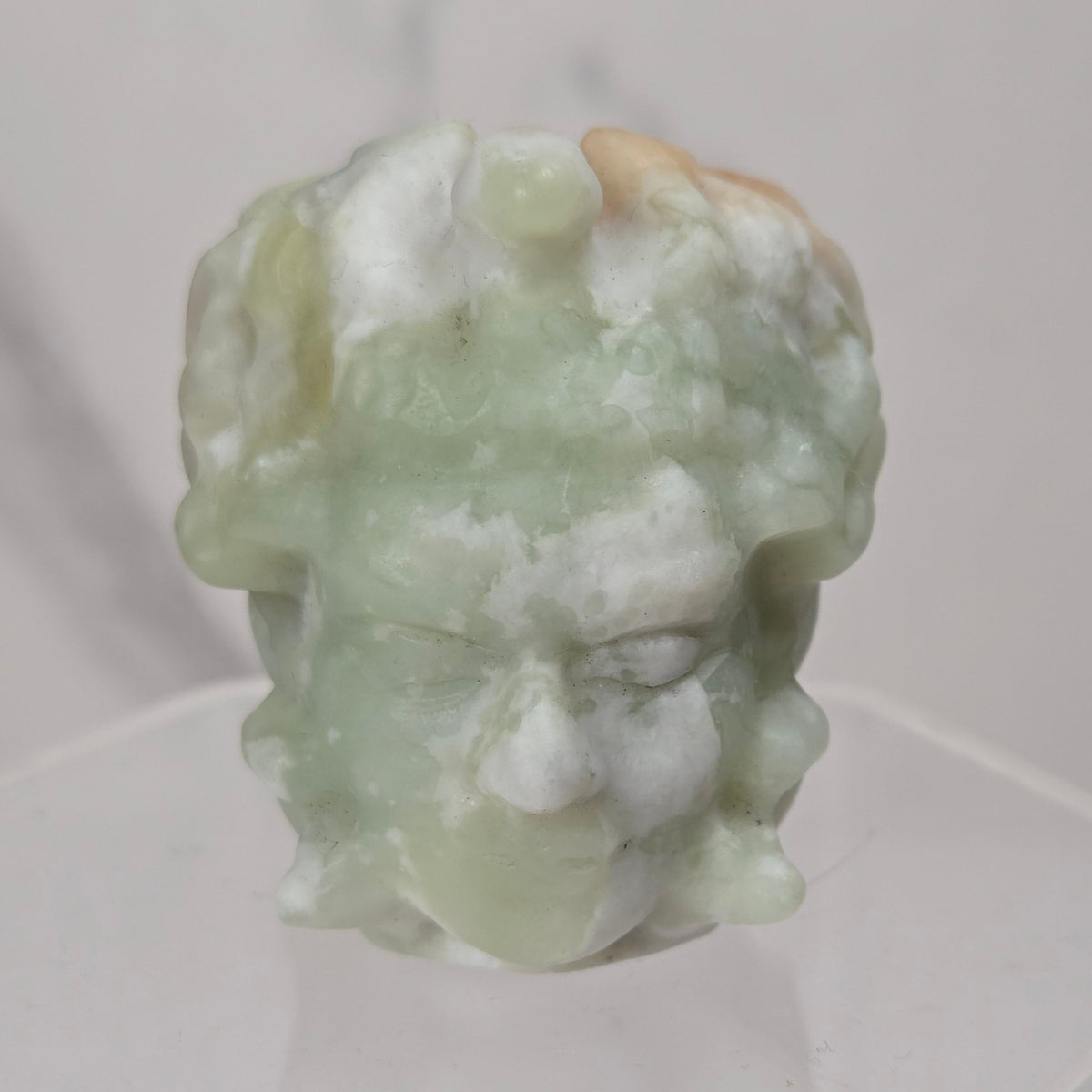 A carved jadeite head sculpture with a light green and white marbled texture, displayed on a clear acrylic pedestal against a light gray marble background.