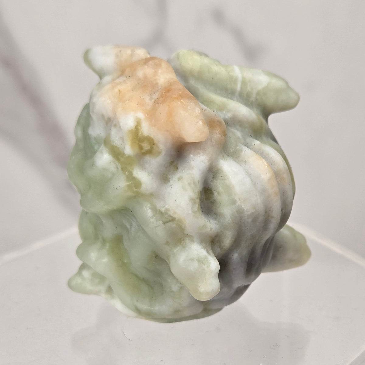 A carved jadeite head sculpture with a light green and white marbled texture, displayed on a clear acrylic pedestal against a light gray marble background.