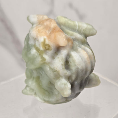 A carved jadeite head sculpture with a light green and white marbled texture, displayed on a clear acrylic pedestal against a light gray marble background.