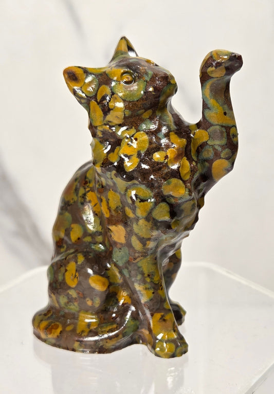 Colorful spotted Ajooba Jasper carving of a cat with it's paw in the air on an acrylic stand against a white background