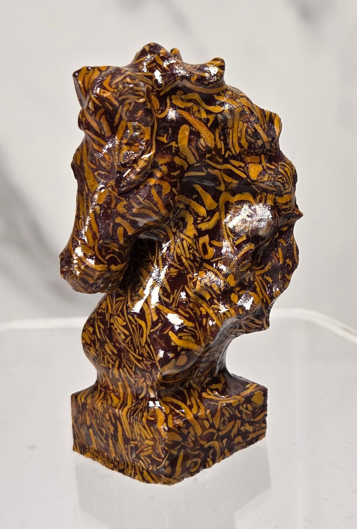 A glossy brown and amber marbled horse head sculpture displayed on a clear acrylic stand against a light gray marble-patterned background.