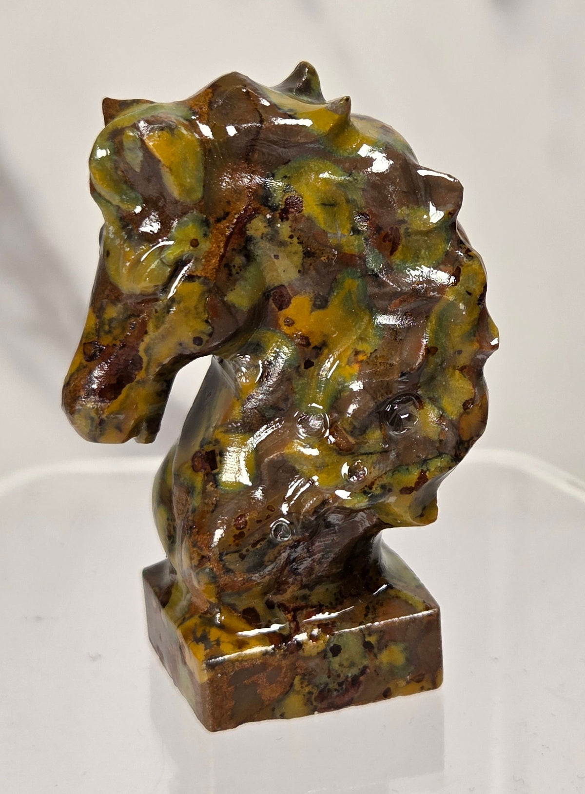A small carved horse head figurine made of a brown and yellow colored stone, displayed on a clear acrylic stand against a light marble-patterned background.