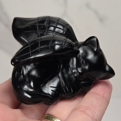 A small black stone figurine of a sleeping cat with wings, held in a person's hand. The figurine has a textured, matte stone finish with gray and white marbling.