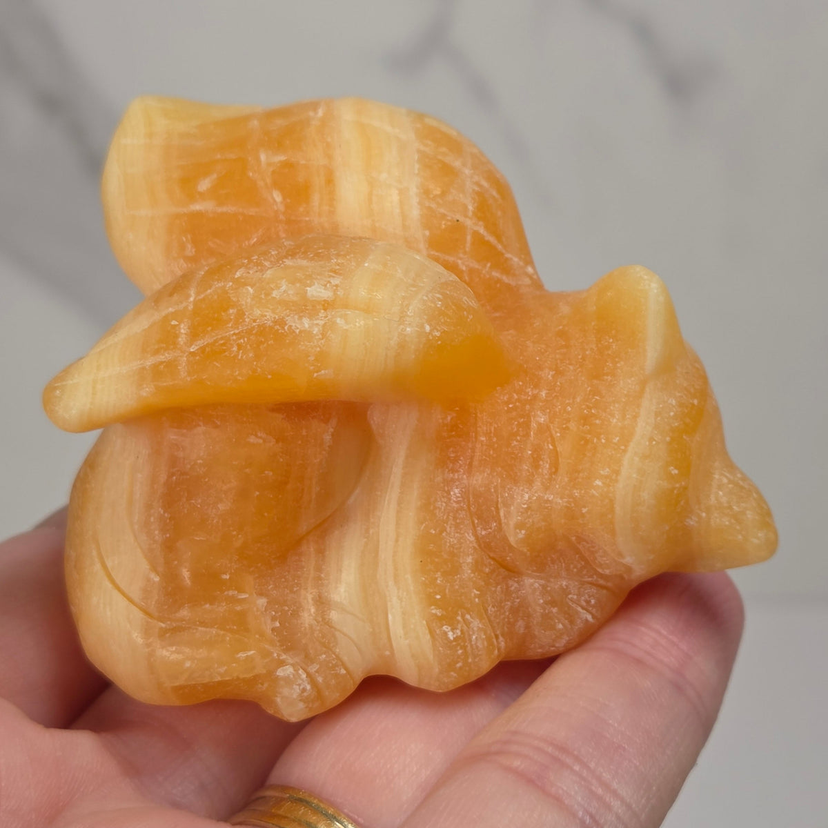 A small orange calcite stone figurine of a sleeping cat with wings, held in a person's hand. The figurine has a textured, matte stone finish with gray and white marbling.