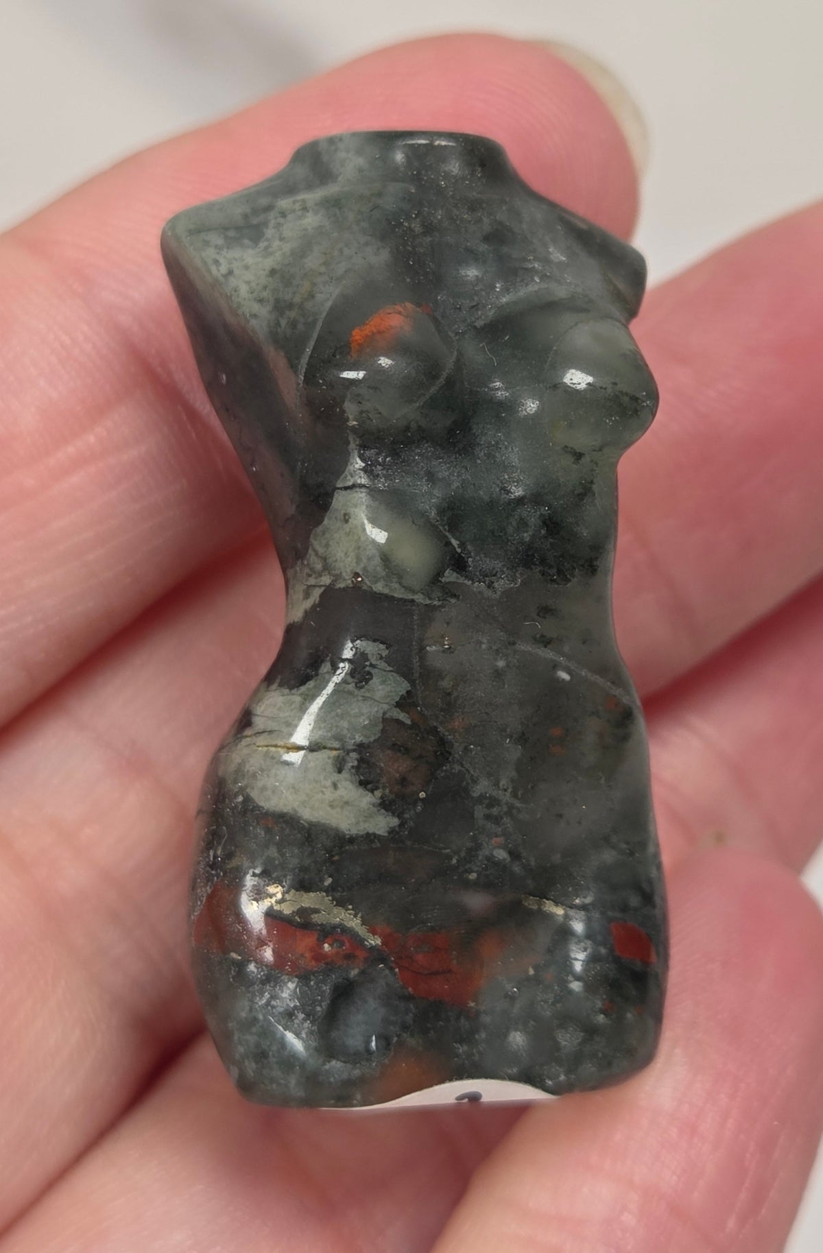A hand holding a small carved stone figurine shaped like a torso, made from  african bloodstone with red, black and grey coloring