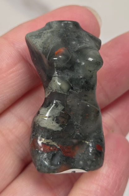 A hand holding a small carved stone figurine shaped like a torso, made from  african bloodstone with red, black and grey coloring