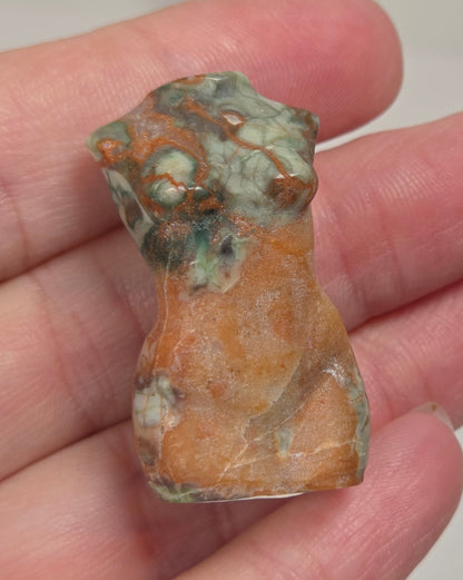 A hand holding a small carved stone figurine shaped like a torso, made from a orange, green and brown colored agate.