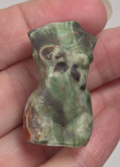 A hand holding a small carved stone figurine shaped like a torso, made from a orange, green and brown colored agate.