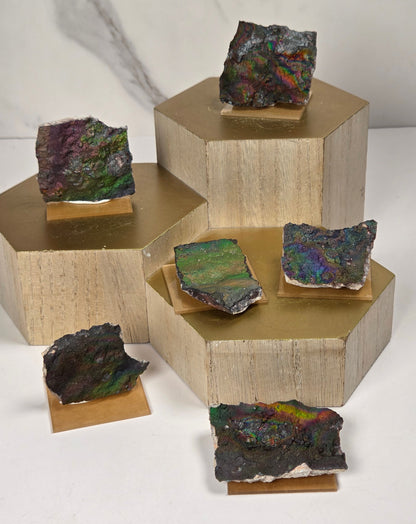 Five raw iridescent mineral or stone slabs with dark charcoal bases and vivid rainbow reflections, arranged on small wooden risers atop a staircase of gold and beige pedestals against a light marble-look background.