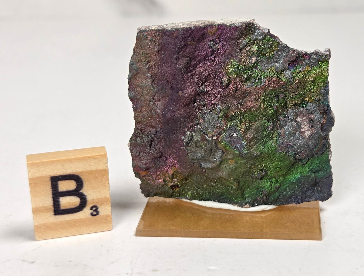 An iridescent mineral or rock specimen with purple, green, and blue tones, displayed on a small tan stand next to a wooden Scrabble tile showing the letter B and value 3, on a white marble-like surface.