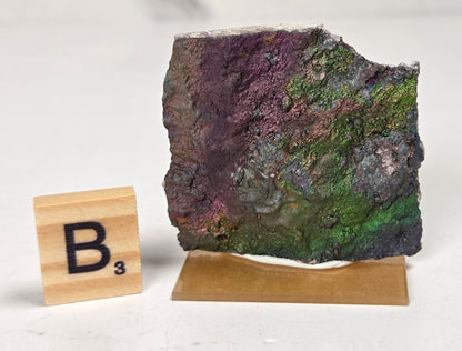 An iridescent mineral or rock specimen with purple, green, and blue tones, displayed on a small tan stand next to a wooden Scrabble tile showing the letter B and value 3, on a white marble-like surface.