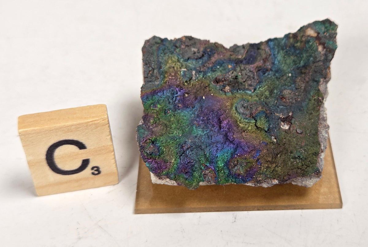 An iridescent mineral or rock specimen with purple, green, and blue tones, displayed on a small tan stand next to a wooden Scrabble tile showing the letter C and value 3, on a white marble-like surface.