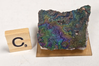 An iridescent mineral or rock specimen with purple, green, and blue tones, displayed on a small tan stand next to a wooden Scrabble tile showing the letter C and value 3, on a white marble-like surface.