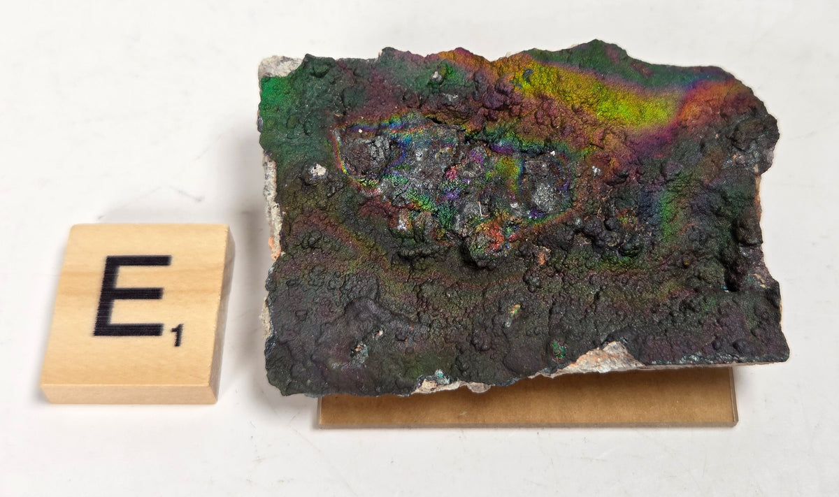 An iridescent mineral or rock specimen with purple, green, and blue tones, displayed on a small tan stand next to a wooden Scrabble tile showing the letter E and value 3, on a white marble-like surface.