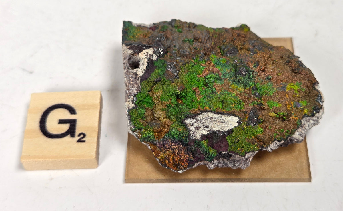 An iridescent mineral or rock specimen with purple, green, and blue tones, displayed on a small tan stand next to a wooden Scrabble tile showing the letter G and value 3, on a white marble-like surface.