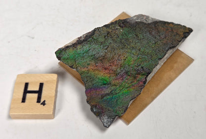 An iridescent mineral or rock specimen with purple, green, and blue tones, displayed on a small tan stand next to a wooden Scrabble tile showing the letter H and value 3, on a white marble-like surface.