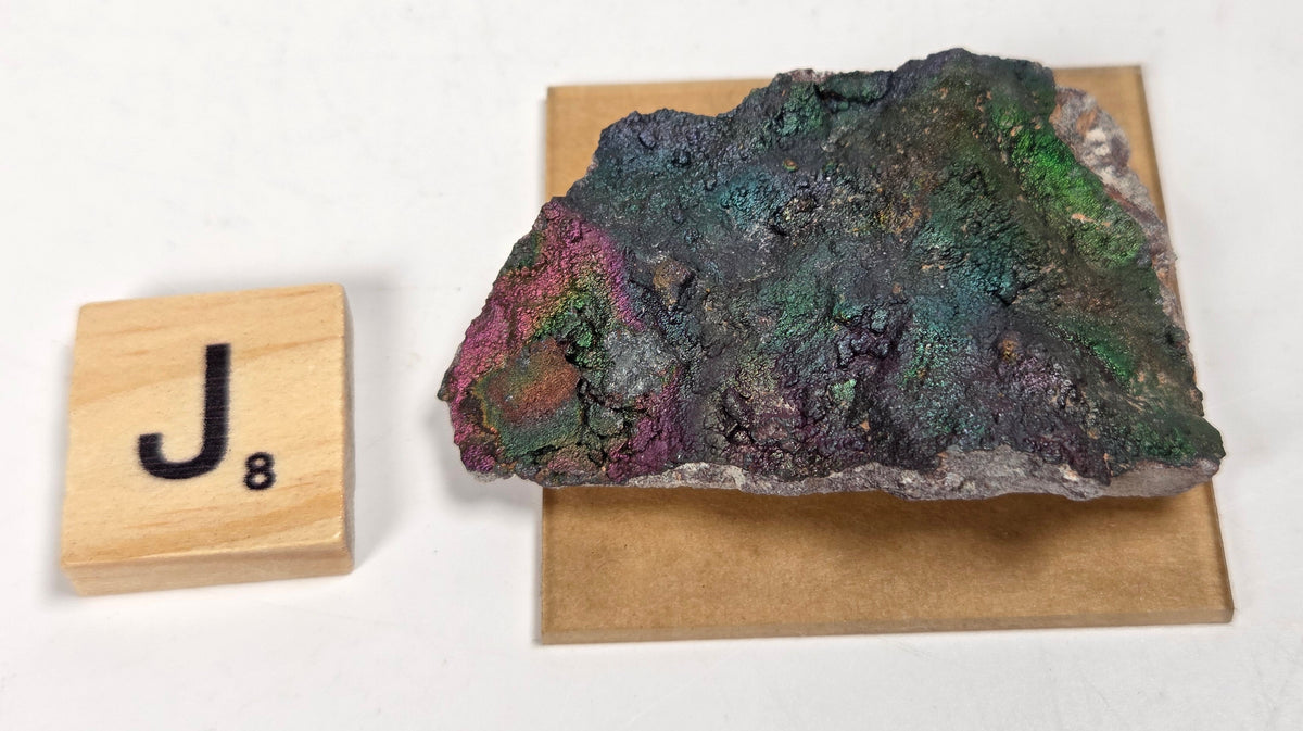 A raw, rough mineral specimen with iridescent green, blue, purple, and pink hues, displayed on a square brown cardboard backing next to a wooden Scrabble tile marked 'J8' on a white surface.