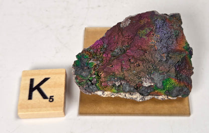 An iridescent mineral or rock specimen with purple, green, and blue tones, displayed on a small tan stand next to a wooden Scrabble tile showing the letter K and value 3, on a white marble-like surface.