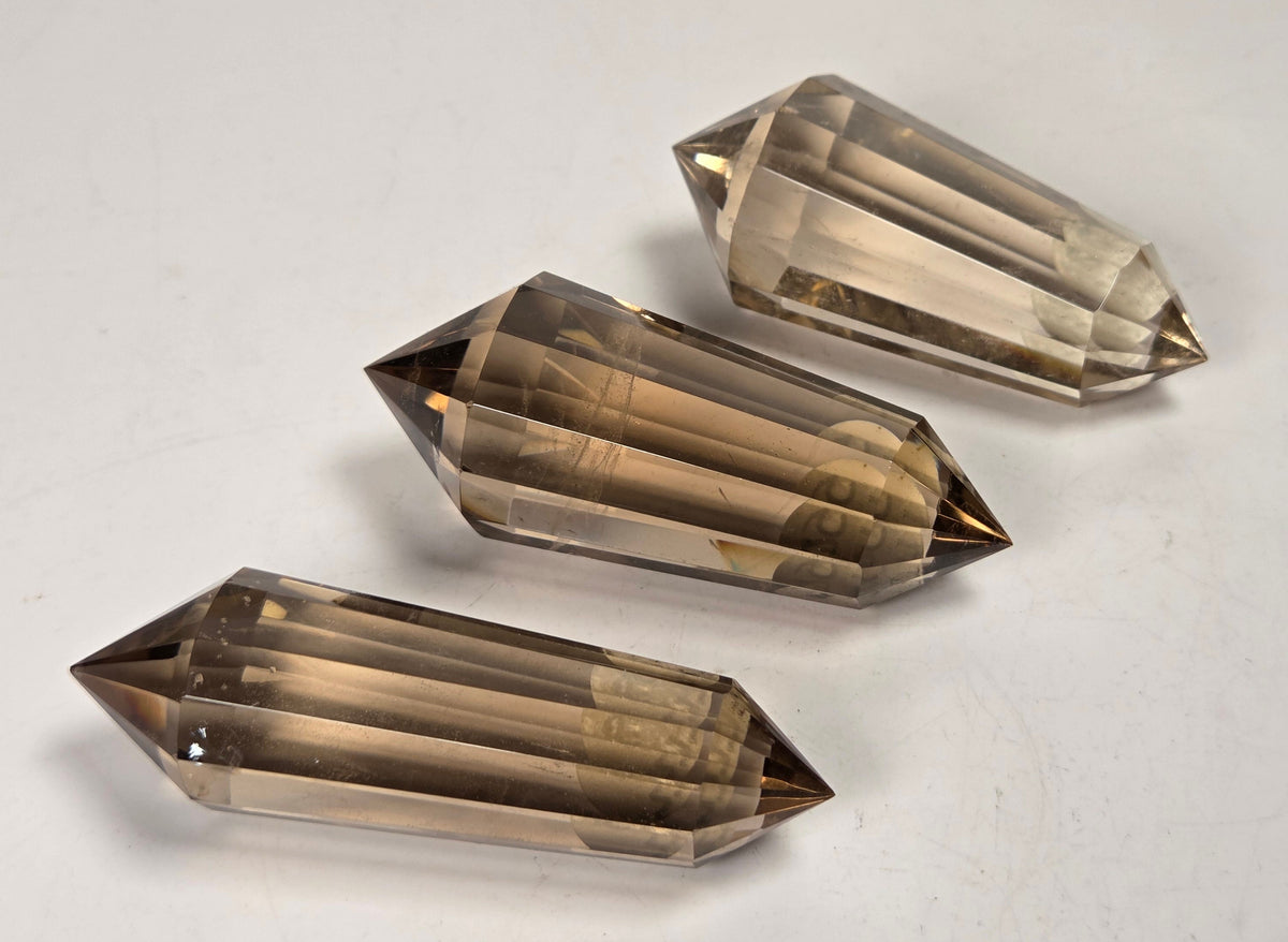 Three faceted smoky quartz crystal points arranged diagonally on a plain off-white surface.