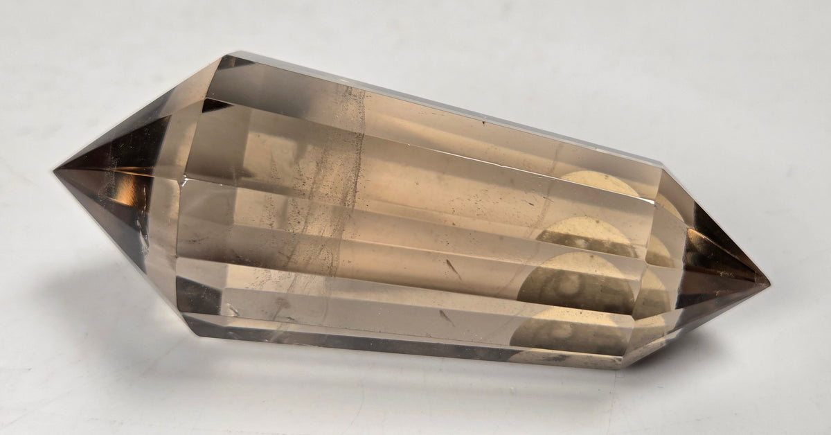 A faceted smoky quartz crystal point, showing a translucent light brown to dark brown gradient, photographed on a neutral light gray surface.