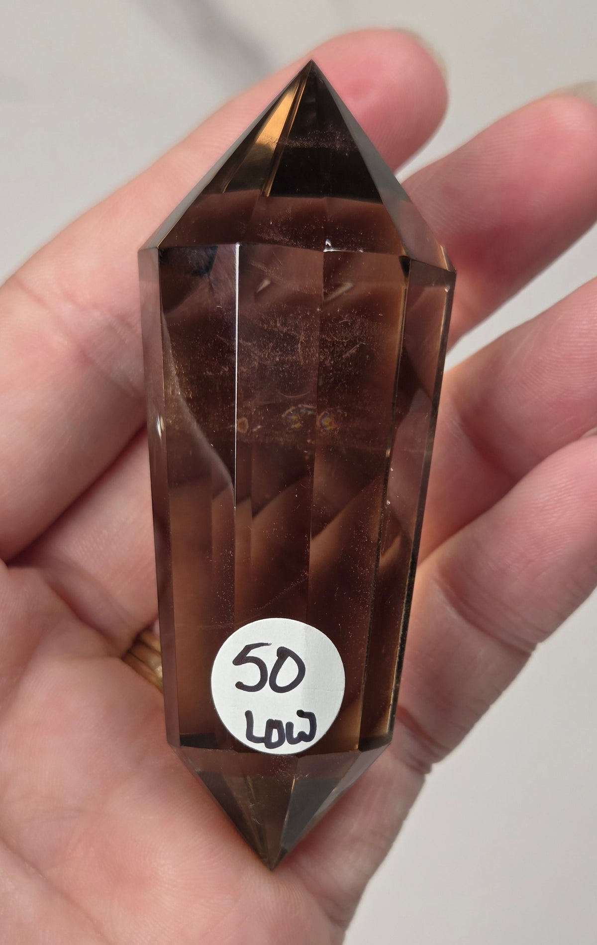 A faceted brown smoky crystal point held in a person's hand, with a small white price sticker reading '50 LDT' attached near the bottom.