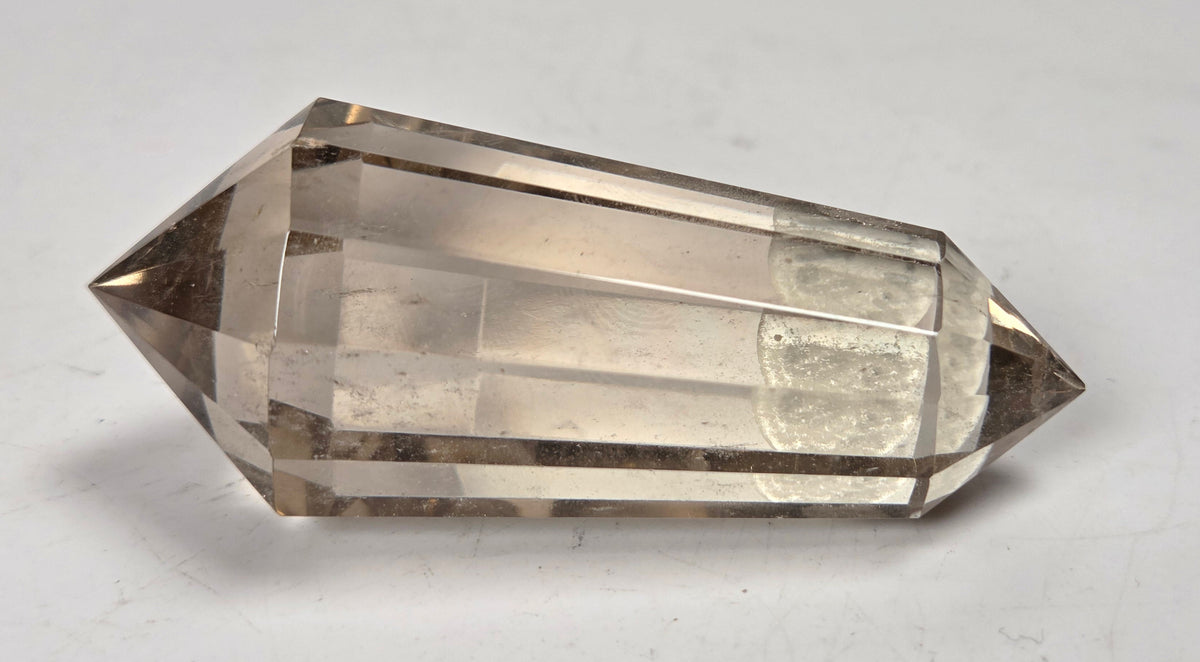 A faceted clear quartz crystal point, also known as a smoky quartz crystal, resting on a plain white surface.