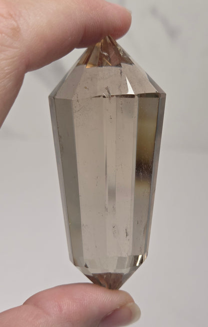 A faceted smoky quartz crystal point held between fingers to show its shape and transparency against a light gray background.