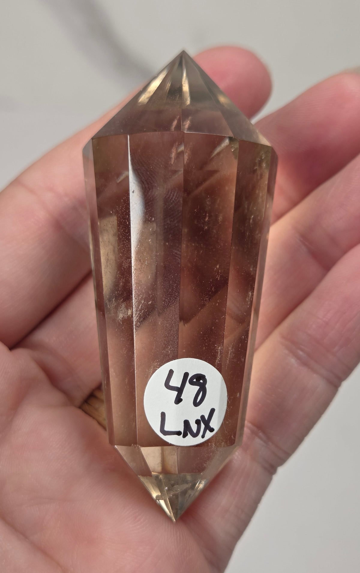 A faceted clear crystal point held in a person's hand against a plain light background. The crystal is transparent with warm brownish-tan internal tones, and a small white sticker with handwritten text is attached near the lower middle.
