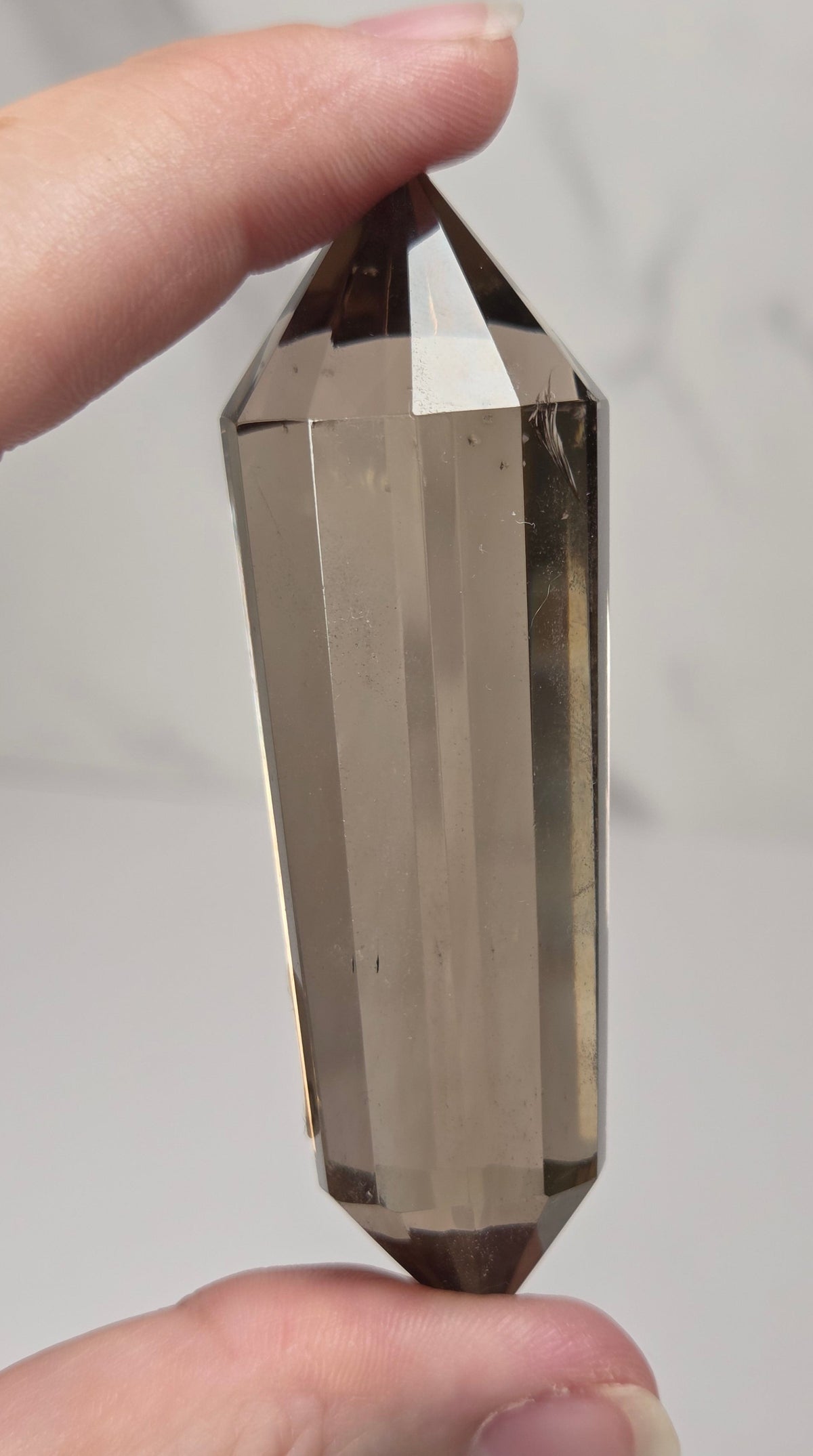 A hand holding a faceted smoky quartz crystal point against a light gray marble background.