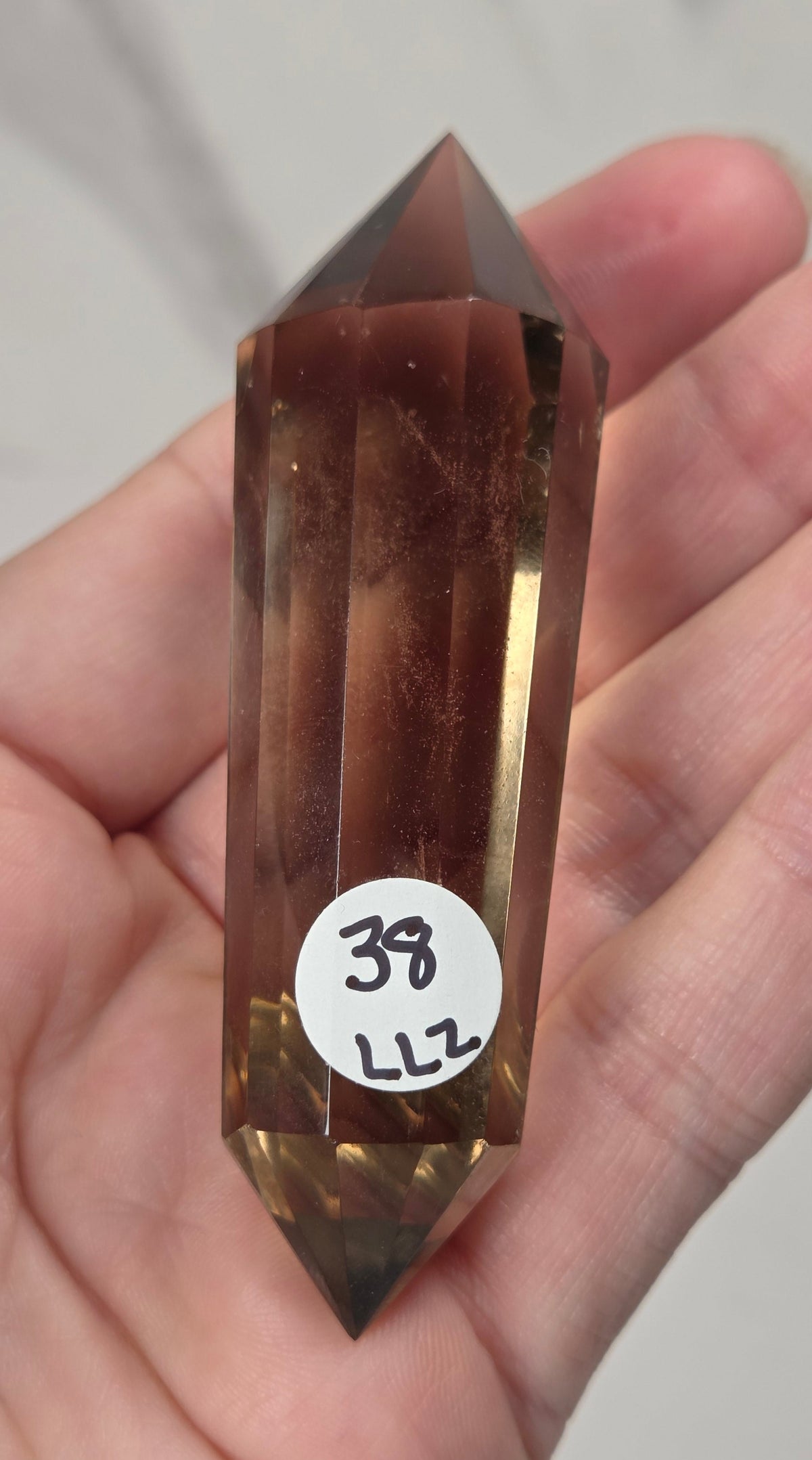 A faceted brown smoky quartz crystal point held in a person's hand, with a small white circular sticker marked '38 L12' attached near the bottom.