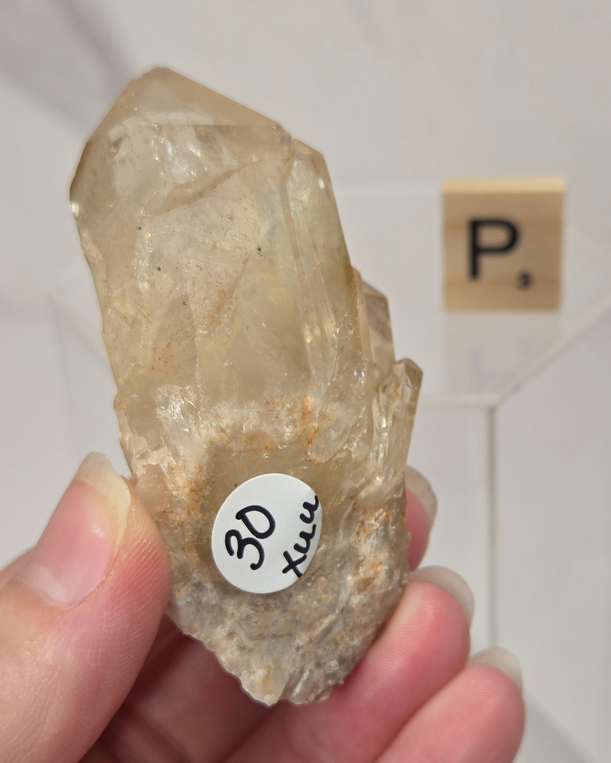 A hand holding a raw, semi-translucent beige quartz crystal cluster, with a small white price tag reading '30 hrs' attached. In the background, a Scrabble tile with the letter 'P' sits on a clear display stand against a light gray marble-patterned wall.