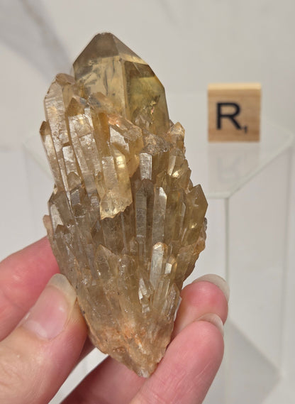 A hand holding a natural quartz crystal point specimen with a rough matrix base. The crystal is translucent golden-brown with some darker inclusions.
