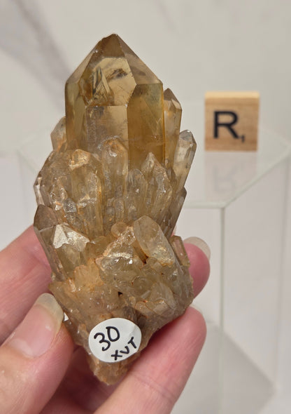 A hand holding a natural smoky quartz crystal cluster with a small white sticker label, set against a light marble background with a Scrabble tile.