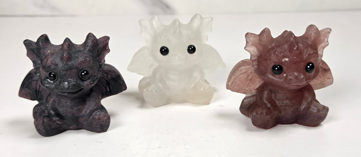 Collection of crystal dragon figurines with large black eyes, sitting on a clear acrylic display stand against a light marble-patterned background.