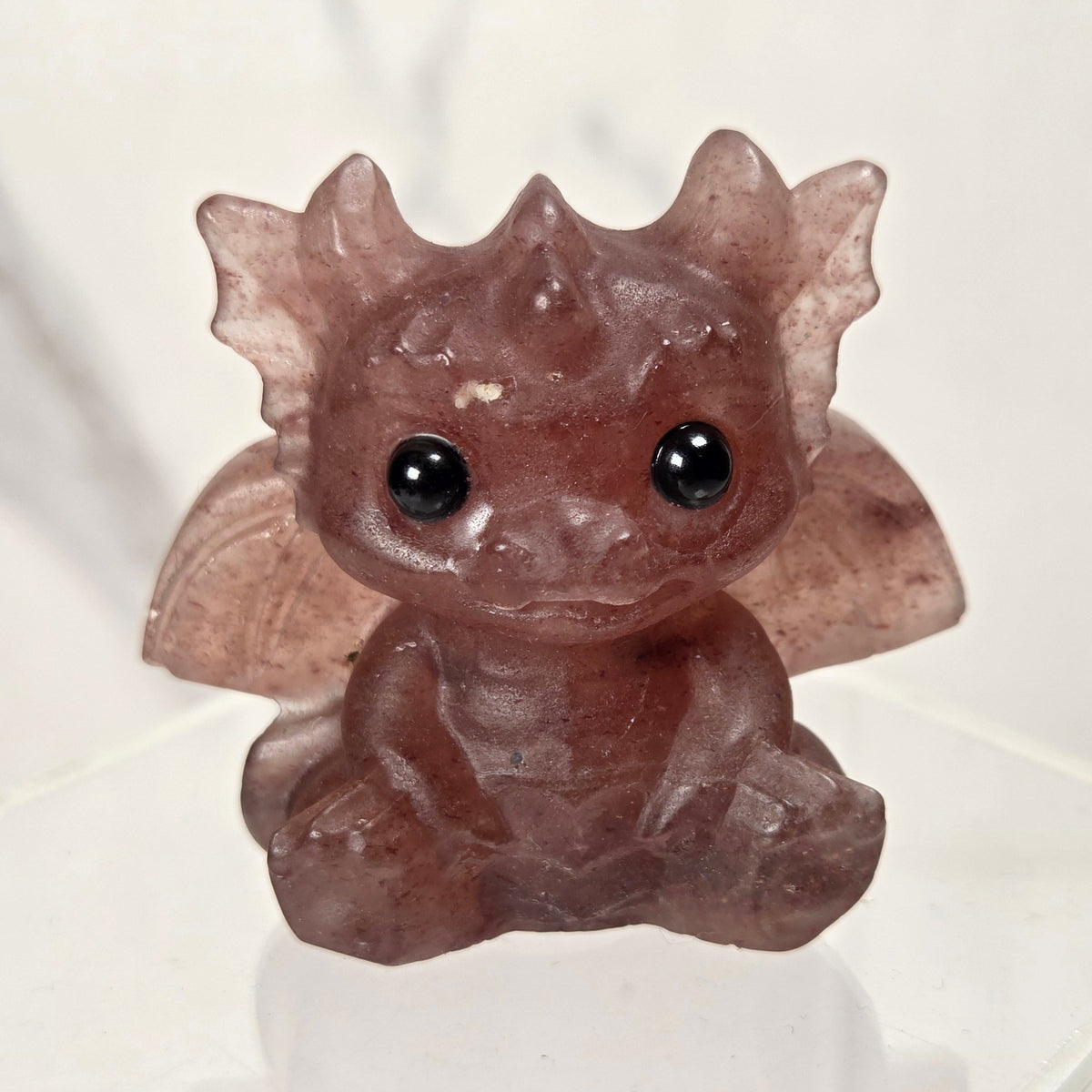A small translucent brown crystal dragon figurine with large black eyes, sitting on a clear acrylic display stand against a light marble-patterned background.